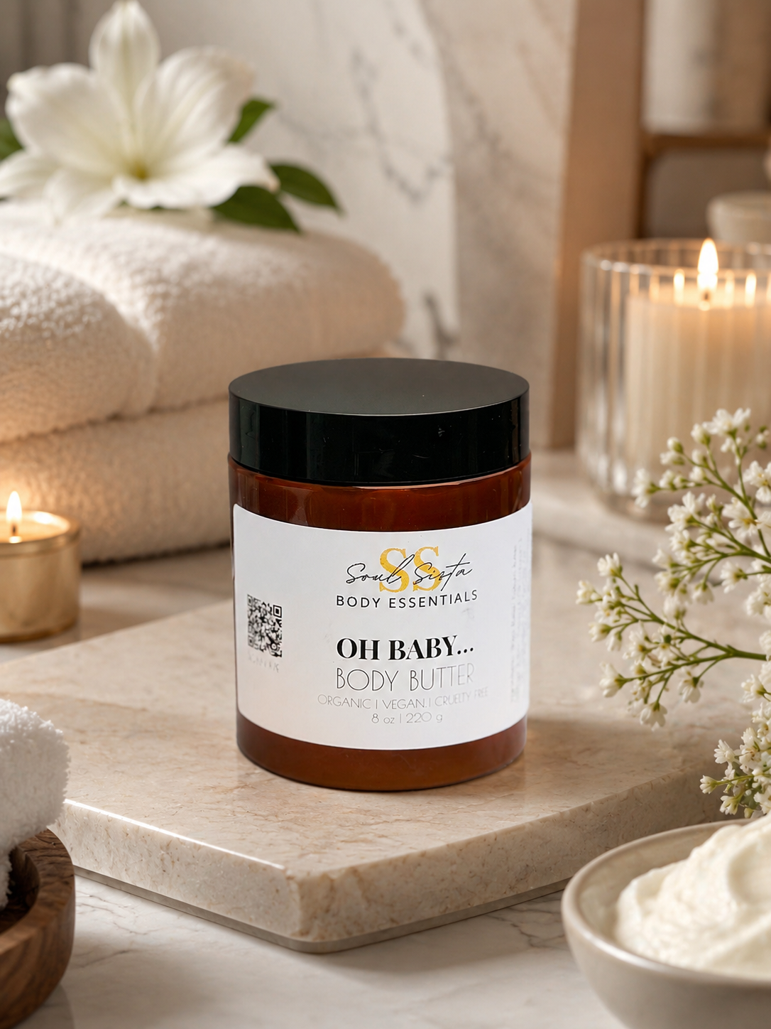 Brown jar of Soulsista Body Essentials Oh Baby Body Butter on a marble surface with candles, flowers, and towels in the background.
